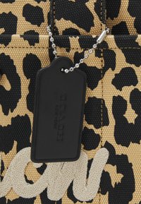 Luipaardprintstof in bruin en zwart met een textuur zwart label bevestigd met een zilveren ketting; bevat een witte geborduurde tekstdetail.