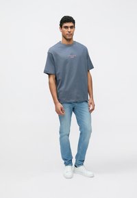 Tricou gri supradimensionat cu guler rotund și logo brodat roz, asortat cu blugi deschis la culoare și adidași albi pe un fundal simplu.