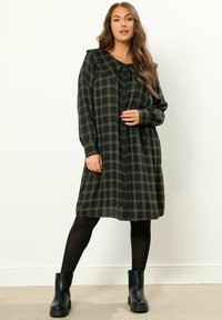 Schwarzes und grünes Karokleid mit langen Ärmeln, großem Kragen und Knopfleiste. Getragen mit schwarzen Knöchelstiefeln und schwarzen Strumpfhosen.