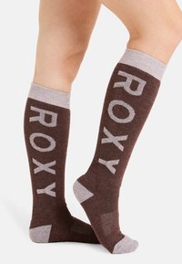 Brązowe skarpetki do kolan z jasnoszarym napisem „ROXY”. Posiadają ribbowaną teksturę oraz kontrastujące jasnoszare akcenty w okolicy mankietu i pięty.