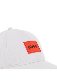 Λευκό καπέλο μπέιζμπολ με ορθογώνιο πορτοκαλί λογότυπο με την επιγραφή «HUGO» σε μαύρο. Το ύφασμα έχει υφή και η γείσος είναι καμπυλωτή.