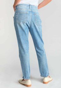 Jean en denim bleu clair avec une coupe décontractée, cinq poches et de subtils détails de couture. Portés avec des mocassins de couleur claire.