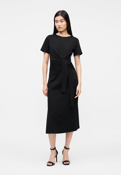Γυναίκα φορώντας μαύρο midi φόρεμα με κοντά μανίκια, λεπτομέρεια δέσιμο στη μέση μπροστά και μαύρα πέδιλα με τακούνι και λουράκι στον αστράγαλο, στέκεται μπροστά σε λευκό φόντο.