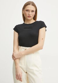 Camiseta negra ajustada de manga corta, con un parche rectangular texturizado en el pecho. Combinada con pantalones crema de talle alto.