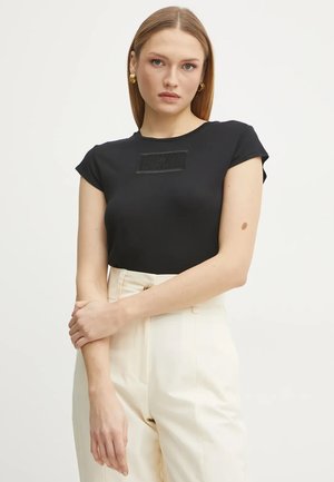 T-shirt nero aderente con maniche corte, caratterizzato da una patch rettangolare testurizzata sul petto. Abbinato a pantaloni beige a vita alta.