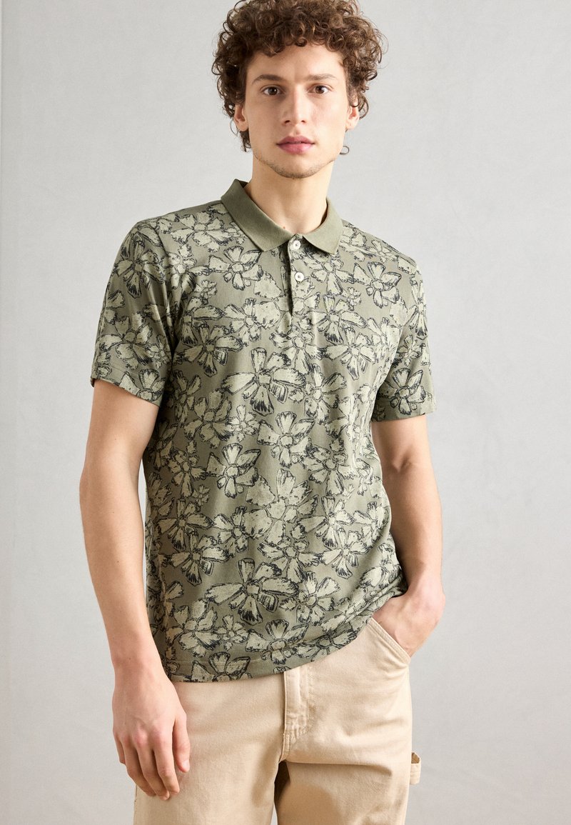 Grünes Poloshirt mit floralem Muster, Kragen, kurzen Ärmeln und einer Knopfleiste mit drei Knöpfen, kombiniert mit hellbeigen Hosen.