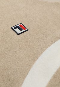 Surface en tissu beige avec un petit patch logo géométrique noir, rouge et blanc, et une bande blanche incurvée partielle.