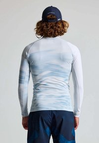 Langärmliger, hellblauer Rashguard mit Wellenmuster, aus glattem, dehnbarem Stoff, getragen mit dunkelgemusterten Shorts und einer marineblauen Mütze.