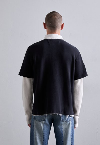 Schwarzes kurzärmliges Oberteil mit strukturierter Oberfläche, über einem weißen langärmligen Hemd getragen, kombiniert mit hellblauen, ausgewaschenen Jeans.