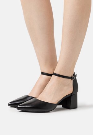 Pieds de femme portant des escarpins à talons carrés pointus en cuir noir avec des sangles autour de la cheville sur un fond blanc.