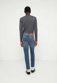 Plave traperice ravnog kroja, s pet džepova i smešnim kožnim detaljem na stražnjem dijelu struka. Siva majica dugih rukava nosi se ispod.