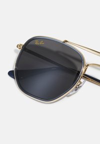 Ray-Ban sončna očala z zlatim kovinskim okvirjem, temnosivimi lečami in kvadratno obliko. Značilnosti vključujejo prozorne nosne blazinice in zlat logotip blagovne znamke.