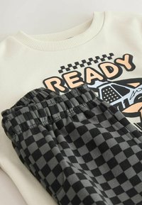 Bluză pulover crem deschis cu imprimeu "READY" și pantaloni jogger în carouri negru-gri, cu talie elastică și textură moale a materialului.