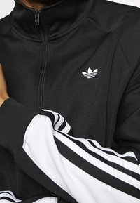 Zwarte zip-up trainingsjack met hoge kraag, witte drie-strips accentueert op de mouwen en een klein wit logo met drie bladeren op de borst.