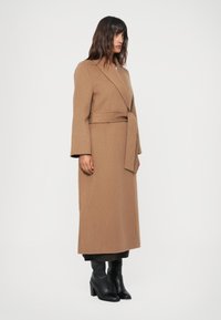 Manteau en laine brun long avec un design enveloppant, des manches larges et une ceinture assortie. Stylisé avec des bottes noires à talons jusqu'aux genoux.