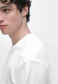 Weißes Kurzarm-T-Shirt mit rundem Ausschnitt, mit glatter Textur und figurbetontem Schnitt, angezeigt am Schulter- und Halsprofil eines Models.