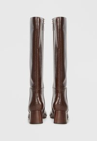 Bottes brunes jusqu'au genou avec un motif reptilien texturé, dotées d'un talon empilé et d'une fermeture éclair sur le côté. Fini en cuir lisse.