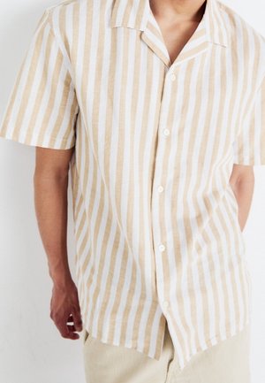 Homme portant une chemise beige et blanche à rayures verticales, manches courtes, col décontracté et pantalon clair.