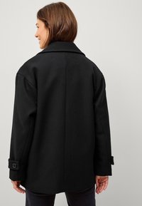 Zwarte wolmix jas met een op maat gemaakt ontwerp, grote kraag, knoopmanchetten en een rechte snit, gezien vanaf de achterkant.