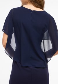 Femme portant une robe bleu marine avec des manches légères et transparentes, vue de dos, mettant en valeur la fluidité du tissu et la fermeture éclair.