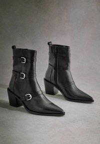 Botines de cuero negro hasta el tobillo con punta afilada, tacón apilado y dos hebillas en los costados. Textura suave con cierre de cremallera en la parte posterior.