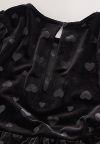 Abito in velluto nero con motivo di piccoli cuori delicati. Schiena con apertura a occhiello chiusa da un singolo bottone. Tessuto morbido e vita arricciata.