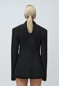 Femeie blondă cu coada de cal, purtând o jachetă neagră supradimensionată și pantaloni scurți negri, arătată din spate pe un fundal simplu gri deschis.