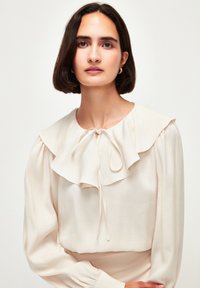 Cremefarvet bluse med et lagdelt, flæset krave, lange ærmer og et bånd ved halsudskæringen. Glat, tekstureret stof.