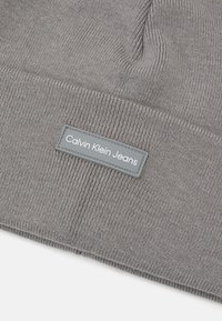 Siva pletena kapa s preklopom. Ima pravokutnu sivu oznaku s natpisom "Calvin Klein Jeans" u bijelom tekstu. Mekana tekstura.