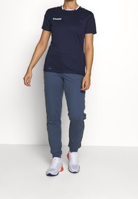 Marinblå tränings-T-shirt med vit kant och logotyp, tillsammans med ljusblå sweatpants. Modellen bär vita sneakers med röda accenter.