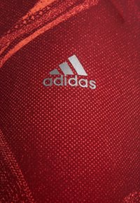 Czerwona koszulka sportowa z teksturowanym wzorem, z logo Adidas w kolorze srebrnym umieszczonym z przodu po lewej stronie.