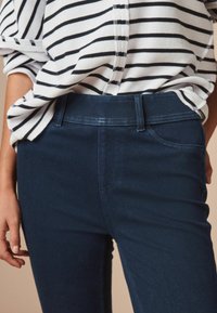 Jeans de mezclilla azul oscuro con una textura suave, que cuentan con una cintura de altura media, bolsillos frontales y costuras limpias. Combinados con una camisa blanca a rayas.