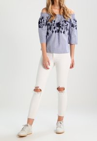 Blouse rayée décolletée, ornée de broderies florales navy, associée à un jean skinny blanc déchiré et des baskets blanches avec des semelles beige clair.