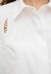 Weiße Bluse mit aufwendiger Stickerei, Perlen und Spitze. Der Button-down-Kragen und die detaillierten Ausschnitte verbessern das Design.