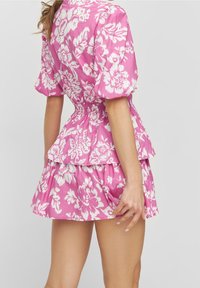 Žena oblečená v krátkých šatech s růžovým a bílým květinovým potiskem, s nabíranými rukávy, elastickým pasem a rozšířenou sukní, viděná zezadu.