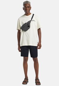 Borsa a tracolla nera con cerniera, di forma rettangolare, indossata sopra una t-shirt crema e pantaloni corti blu navy. Modella che indossa sandali marroni, in posizione verticale.