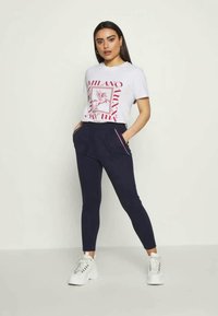 T-shirt branca com estampagem gráfica vermelha e calças de gangue azul-marinho com detalhes cor-de-rosa; modelo a usar ténis brancos, com as mãos nos bolsos.