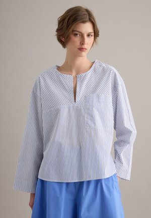 Mujer con una blusa blanca con delgadas rayas verticales azules y una pequeña abertura en V en el cuello, combinada con pantalones azules sueltos, de pie frente a un fondo liso.