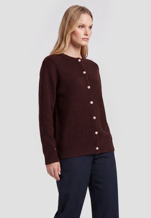 Kvinde iført en mørkebrun knapet cardigan med perleknapper og marineblå bukser, stående foran en ensfarvet lys baggrund.
