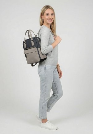 Frau trägt hellgrauen Pullover und Jeans, lächelt, trägt einen zweifarbigen schwarzen und grauen Rucksack auf dem Rücken, steht vor weißem Hintergrund.