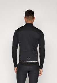 Svart langermet sykkeljakke med glatt tekstur, glidelåslomme bak, reflekterende stripe og elastisk kant for en sikker passform.