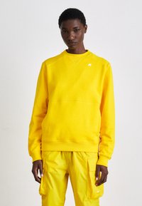 Felpa gialla realizzata in materiale morbido, con colletto rotondo, polsini a coste e un piccolo logo sul petto. Indossata con pantaloni gialli coordinati.