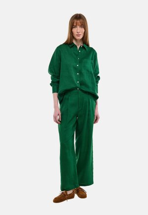 Femme debout, face à nous, portant une chemise verte ample à boutons, un pantalon large assorti et des chaussures marron à lacets, sur un fond uni.