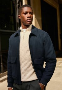 Marineblaues Zip-Jackett mit zwei vorderen Taschen, über einem hellen Pullover mit Rautenmuster getragen, kombiniert mit maßgeschneiderten Hosen.
