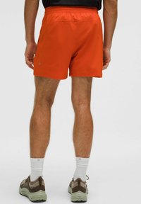 Mann in orange Sportshorts, weißen Socken und braunen Sneakers, der mit sichtbaren Beinen von hinten vor einem schlichten Hintergrund steht.