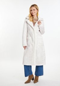 Cappotto bianco in pelliccia sintetica con colletto a scialle e due tasche frontali, indossato sopra un maglione a fantasia e jeans in denim a gamba larga.