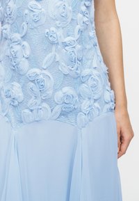 Φωτεινό γαλάζιο φόρεμα με σώμα από ανάγλυφη δαντέλα με λουλουδένιο σχέδιο και αέρινη φούστα από σιφόν, εμφανιζόμενο από τη μέση έως τον βραχίονα με το χέρι να ακουμπάει φυσικά.