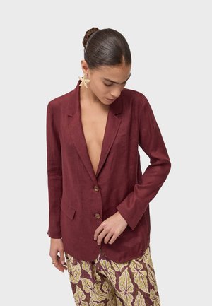 Donna con capelli scuri raccolti in uno chignon che indossa un blazer bordeaux e pantaloni a fantasia, guarda verso il basso, con grandi orecchini a forma di stella.