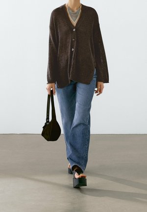 Personne portant un cardigan marron foncé boutonné, un jean bleu, des sandales à plateforme noires, tenant un petit sac à main noir.