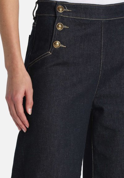 Mørk denimbukser med høj talje, der har tre guldknapper i siden og kontrasterende gul syning langs sømmene.